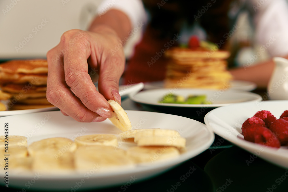 Asian happy skilled pastry housewife in  red apron decorate stack of pancake dessert topping with mixed fruit kiwi raspberry apple from material dish in blurred foreground