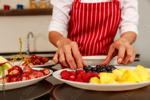 Closeup shot of chef hand putting pieces of fruits into tasty yummy delicious sweet mixed fruit salad in white bowl on kitchen table. Organic and natural health food concept.