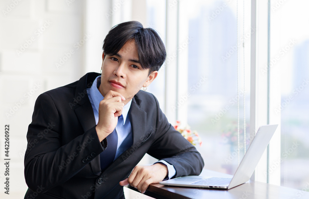 Asian young businessman are smart and handsome wearing black suit sitting while typing on keyboard at laptop near window in office at the morning Asian young businessman are smart and handsome wearing black suit sitting while typing on keyboard at laptop near window in office at the morning