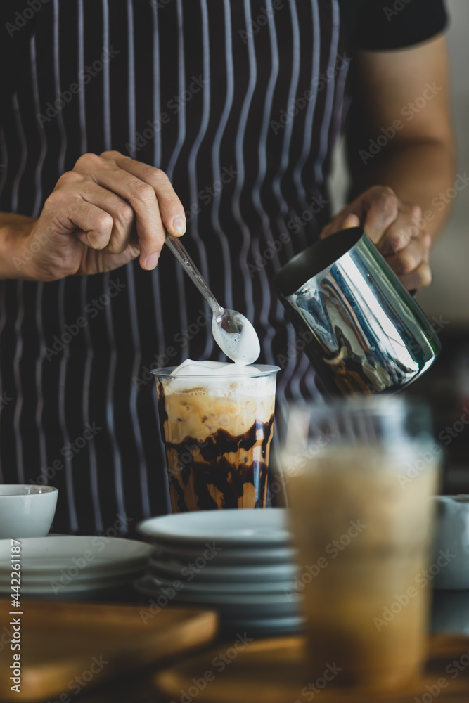Unrecognizable  barista using spoon to gentle topping glass of sweet coffee at bar counter with white milk froth from metal jar to enhance taste of softness and mellowness.