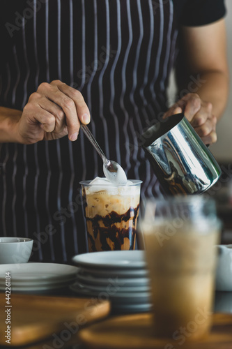 Unrecognizable barista using spoon to gentle topping glass of sweet coffee at bar counter with white milk froth from metal jar to enhance taste of softness and mellowness.