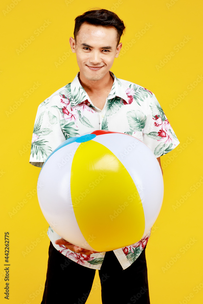 Young attractive Asian man in white Hawaiian shirt holding beach ball against yellow background. Concept for beach vacation holiday Young attractive Asian man in white Hawaiian shirt holding beach ball against yellow background. Concept for beach vacation holiday