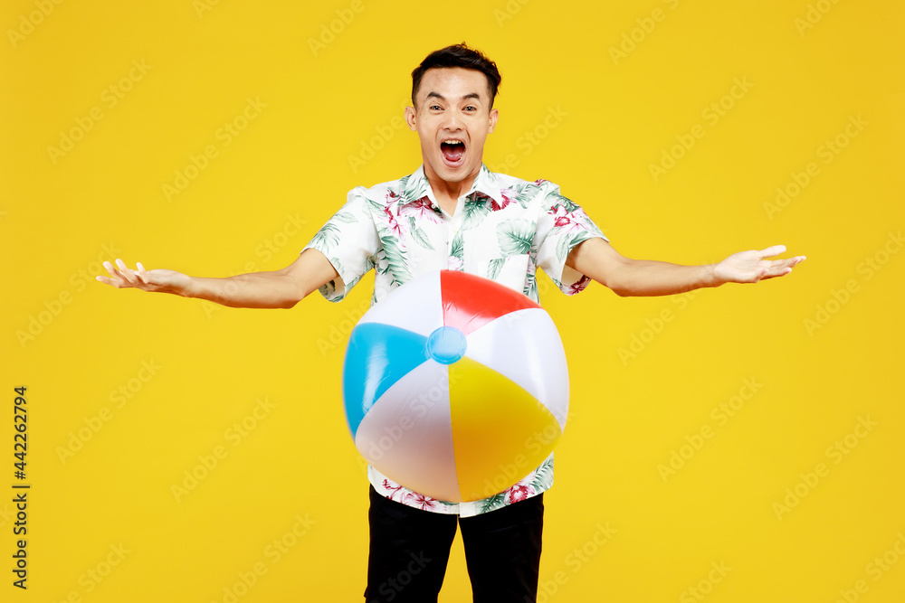 Young attractive Asian man in white Hawaiian shirt holding beach ball against yellow background. Concept for beach vacation holiday