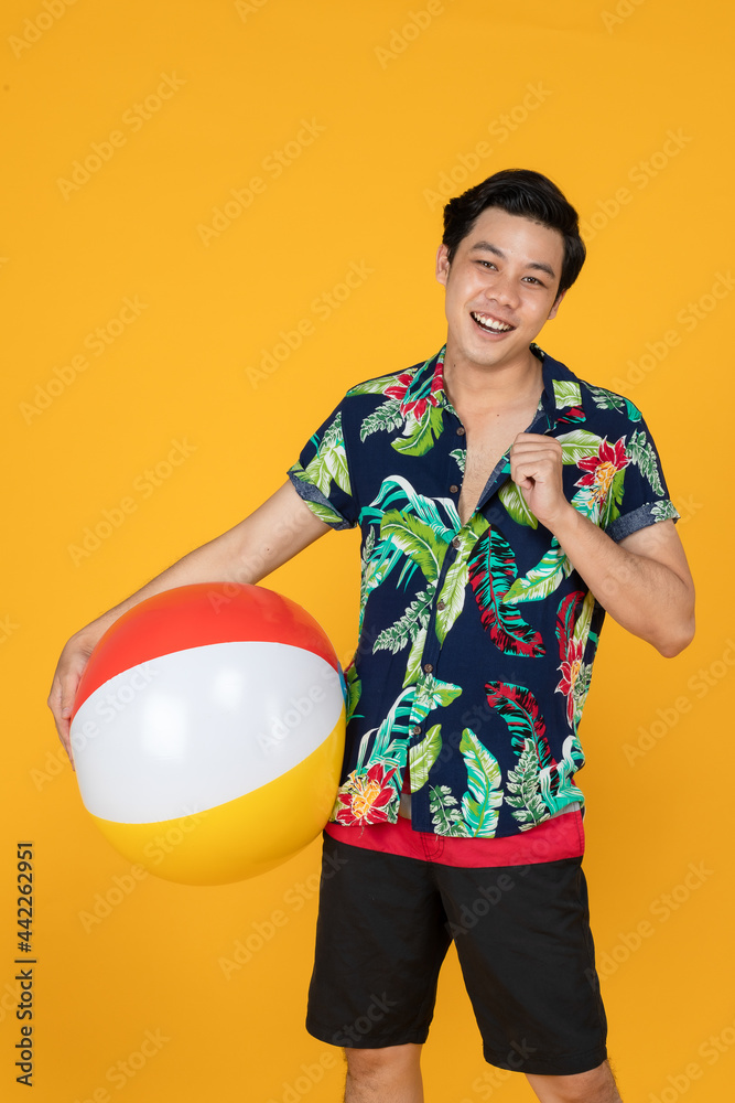 Young attractive Asian man in blue and green Hawaiian shirt holding beach ball against yellow background. Concept for beach vacation holiday Young attractive Asian man in blue and green Hawaiian shirt holding beach ball against yellow background. Concept for beach vacation holiday