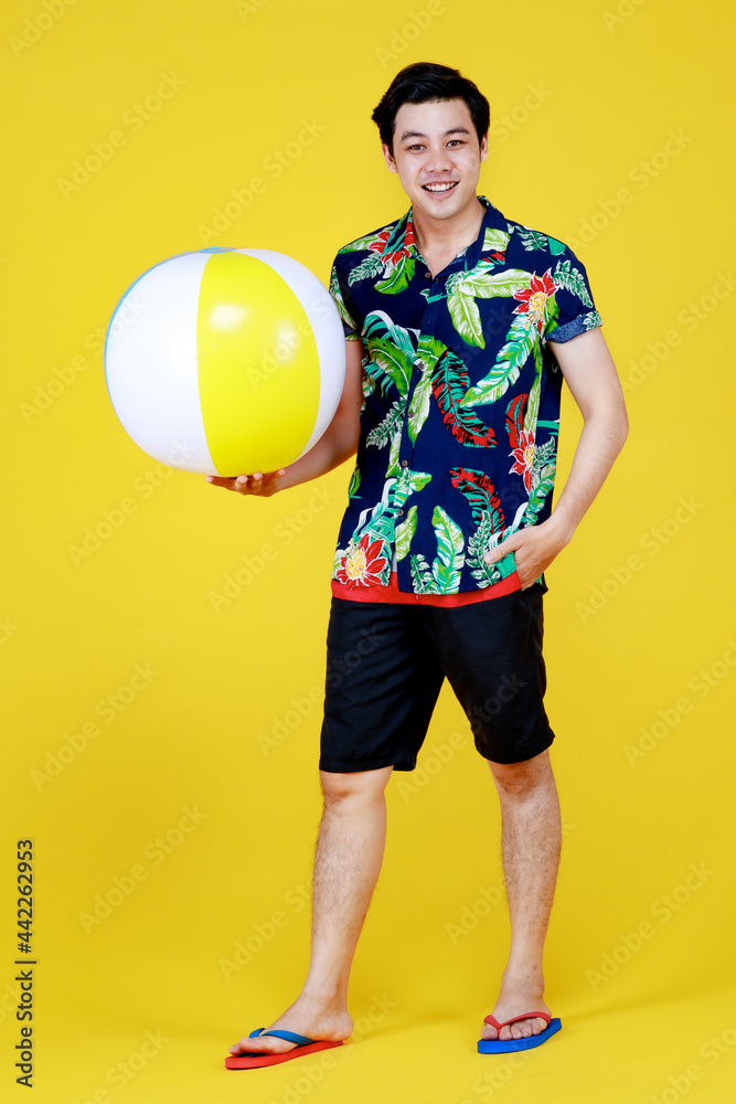 Young attractive Asian man in blue and green Hawaiian shirt holding beach ball against yellow background. Concept for beach vacation holiday