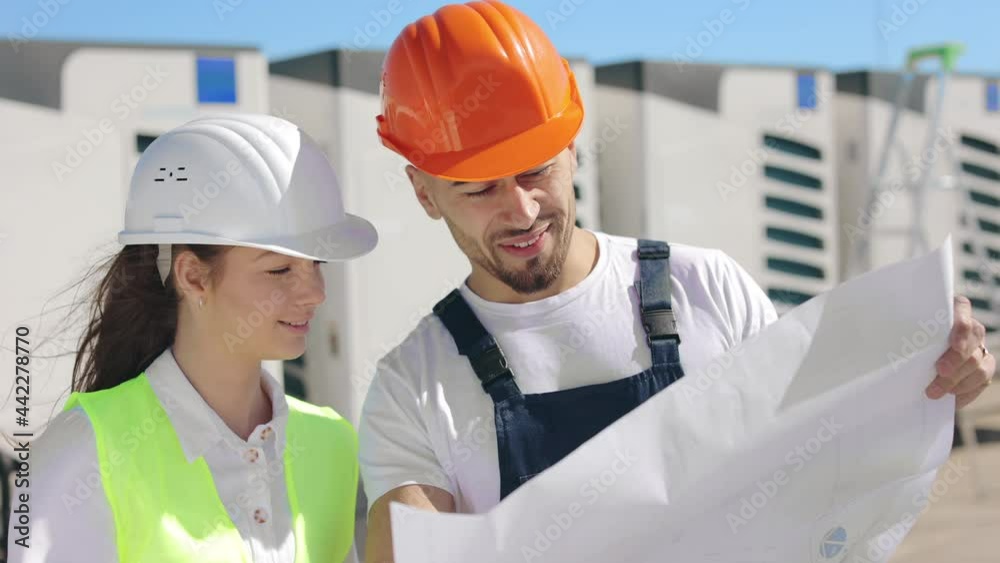 Two happy two engineers are trying to inspect the drawings but the wind is blowing very hard. They are smiling. They are wearing work clothes and a hard hat. Air conditioning system in the background Two happy two engineers are trying to inspect the drawings but the wind is blowing very hard. They are smiling. They are wearing work clothes and a hard hat. Air conditioning system in the background
