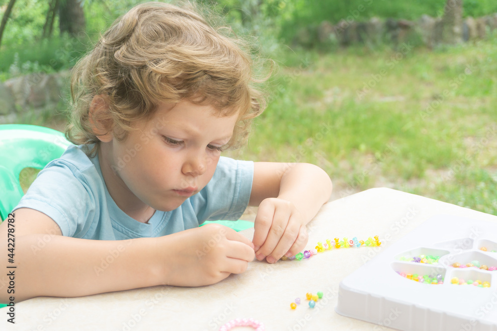 A little Caucasian girl, curly blonde, is captivated by creativity in the fresh air. The girl makes a homemade bead bracelet. A little Caucasian girl, curly blonde, is captivated by creativity in the fresh air. The girl makes a homemade bead bracelet.