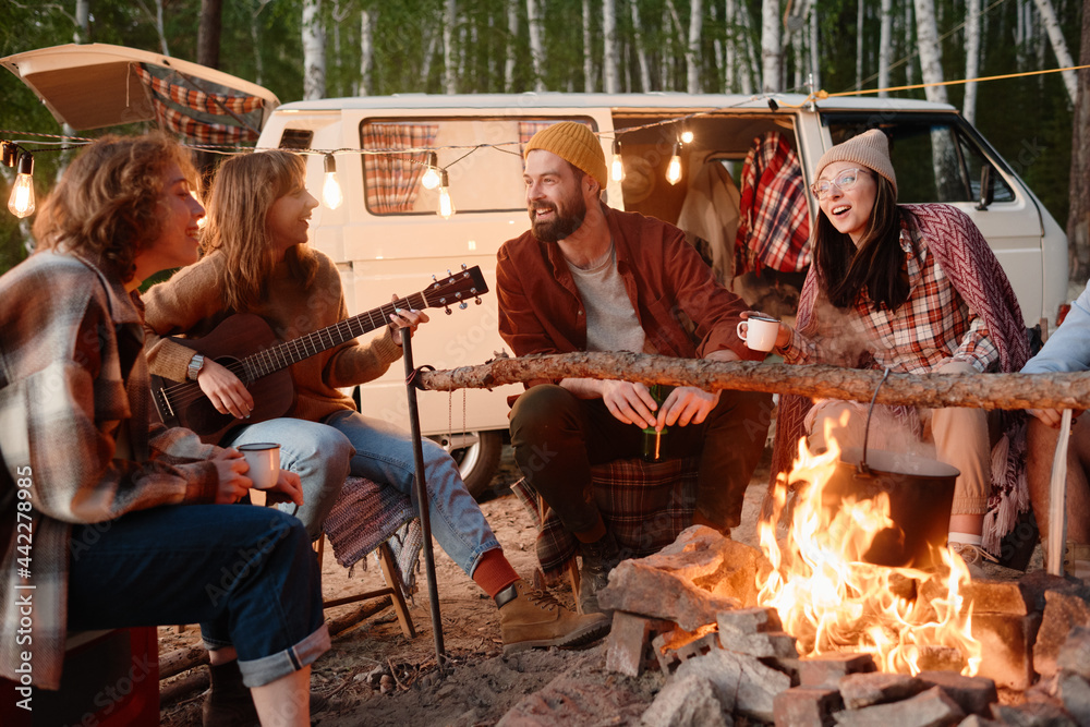 Group of friends sitting near the fire and singing songs with guitar during camping in the forest