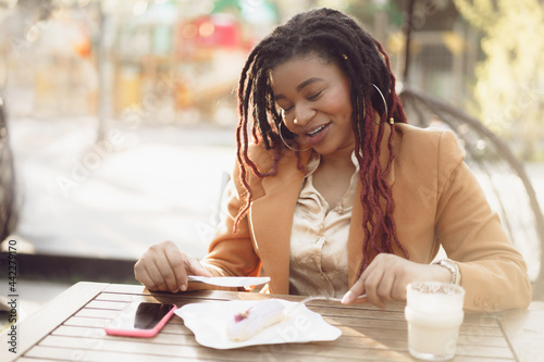 Smiling african american woman drinking coffee and eating dessert in outdoor cafe