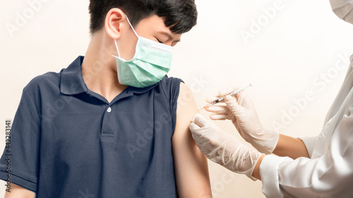 COVID 19 Vaccination for kids and teenager concept. Smart Asian teen boy with medical face mask looking at nurse's hand with syringe, receiving vaccine injection at hospital. Herd immunity, safe, prot