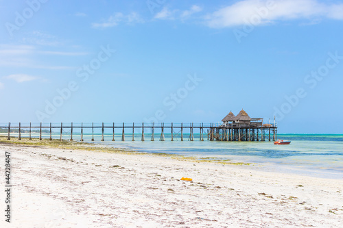 Wooden pier with boat and beach hut on sunny day. Low tide of Indian Ocean. Idyllic exotic resort. Pier in perspective with bungalows. Tropical paradise. Honeymoon in lagoon. Scenic tranquil seascape.
