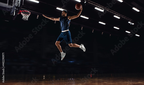 Young African sportsman, basketball player training in gym, idoors isolated on dark background. Concept of sport, game, competition. Slam dunk.