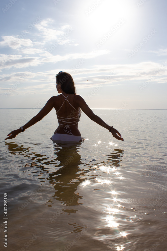 A girl from the back in a white swimsuit enters the sea sparkling from the sun