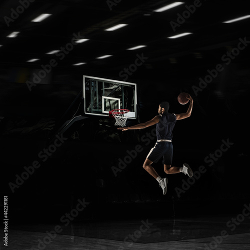 Young African sportsman, basketball player training in gym, idoors isolated on dark background. Concept of sport, game, competition. Slam dunk.