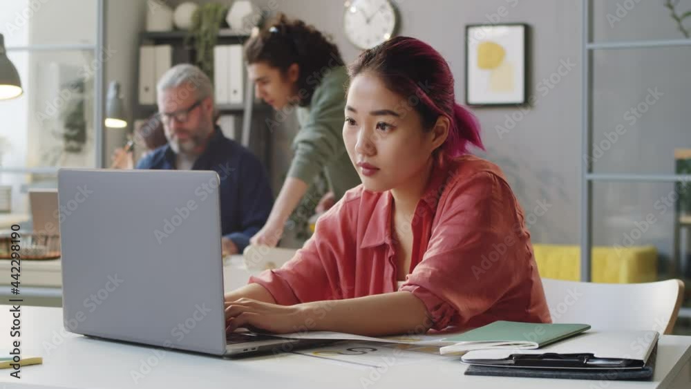 Young Asian female trainee with pink hair working on laptop at desk in office Young Asian female trainee with pink hair working on laptop at desk in office