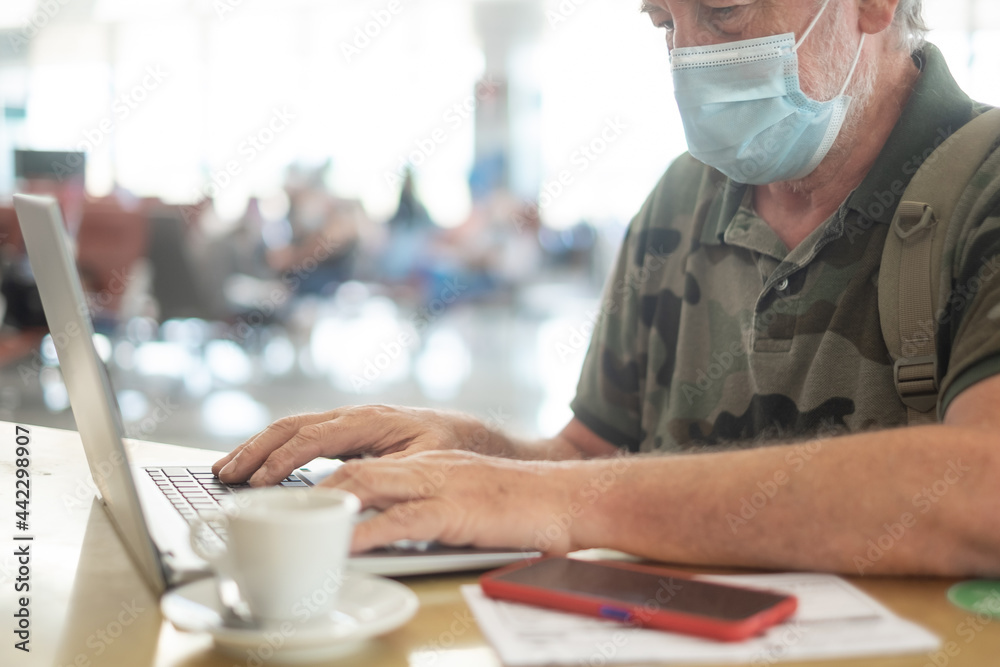Traveler man in airport waiting for boarding works on laptop computer, senior wearing surgical mask due to coronavirus Traveler man in airport waiting for boarding works on laptop computer, senior wearing surgical mask due to coronavirus