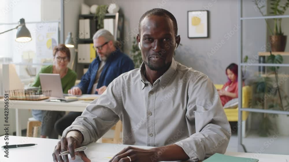 Afro-American man in wireless earphones waving at camera, showing document and explaining details on online video call while working in office Afro-American man in wireless earphones waving at camera, showing document and explaining details on online video call while working in office