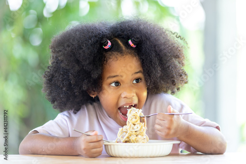 A little curly-haired African American girl sits at the table eating delicious Spaghetti Carbonara. Fun, cheerful. Appetite. Childhood and eating concepts.