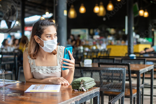 Asian women wearing masks to protect against covid infection Sitting on a smartphone in a restaurant waiting for friends to eat together During the coronavirus outbreak.Concept New Normon .