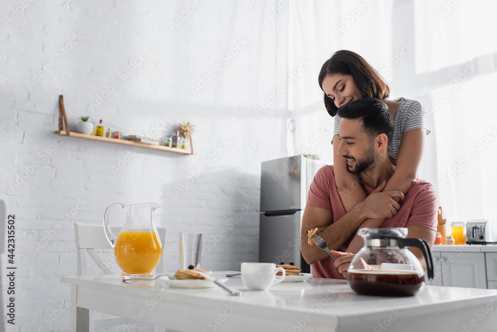smiling young woman gently hugging boyfriend near table with breakfast, orange juice and coffee in kitchen smiling young woman gently hugging boyfriend near table with breakfast, orange juice and coffee in kitchen