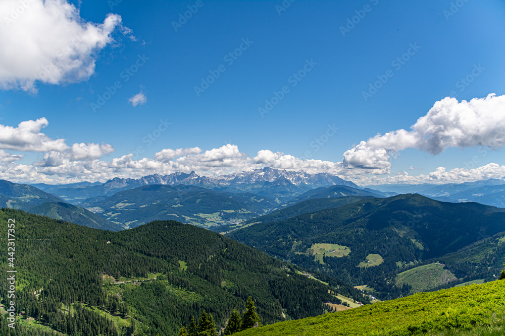 Panoramic view over the Austrian Alps with a hiking trail, trees, and a blue sky with clouds