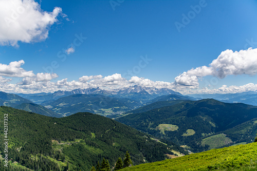 Panoramic view over the Austrian Alps with a hiking trail, trees, and a blue sky with clouds © Lukas