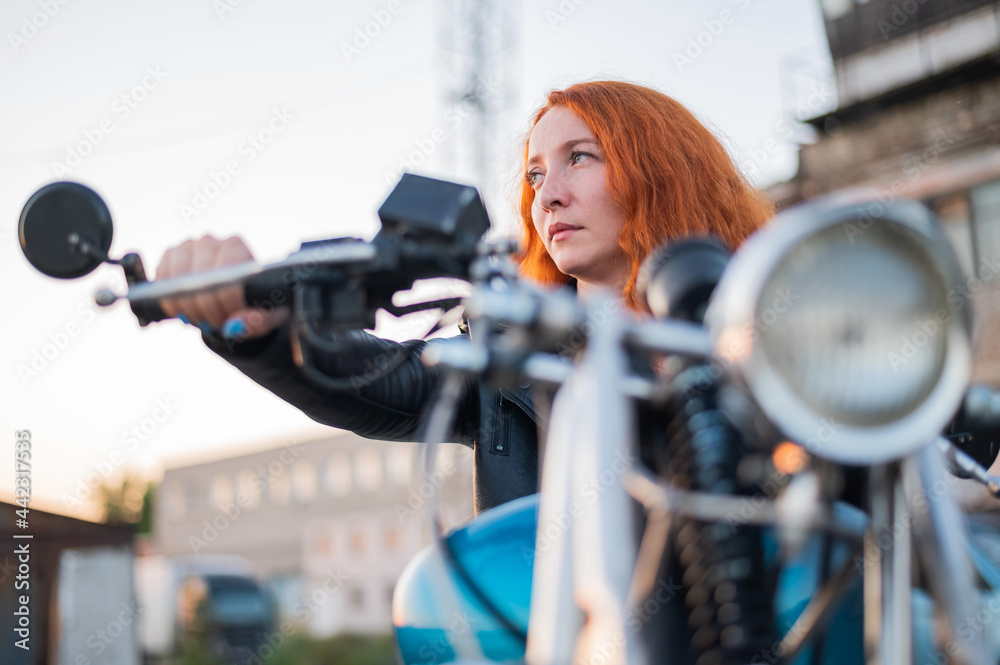 Fototapeta premium Curly red-haired woman in a black leather jacket sits on a motorcycle. Portrait of a serious girl driving a bike.