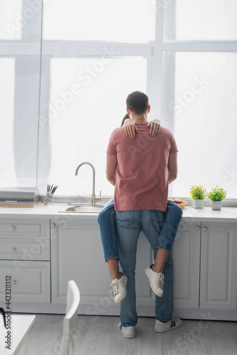 back view of young man hugging girlfriend sitting on cupboard in kitchen