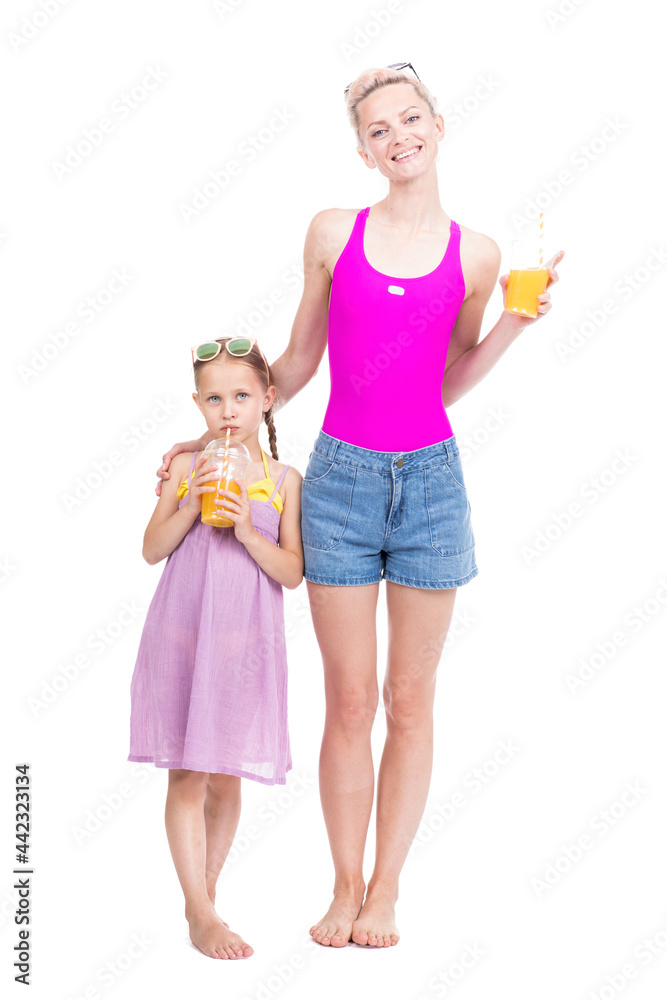 Vertical full length studio portrait of modern Caucasian woman and girl wearing summer clothes standing together drinking fresh orange juice