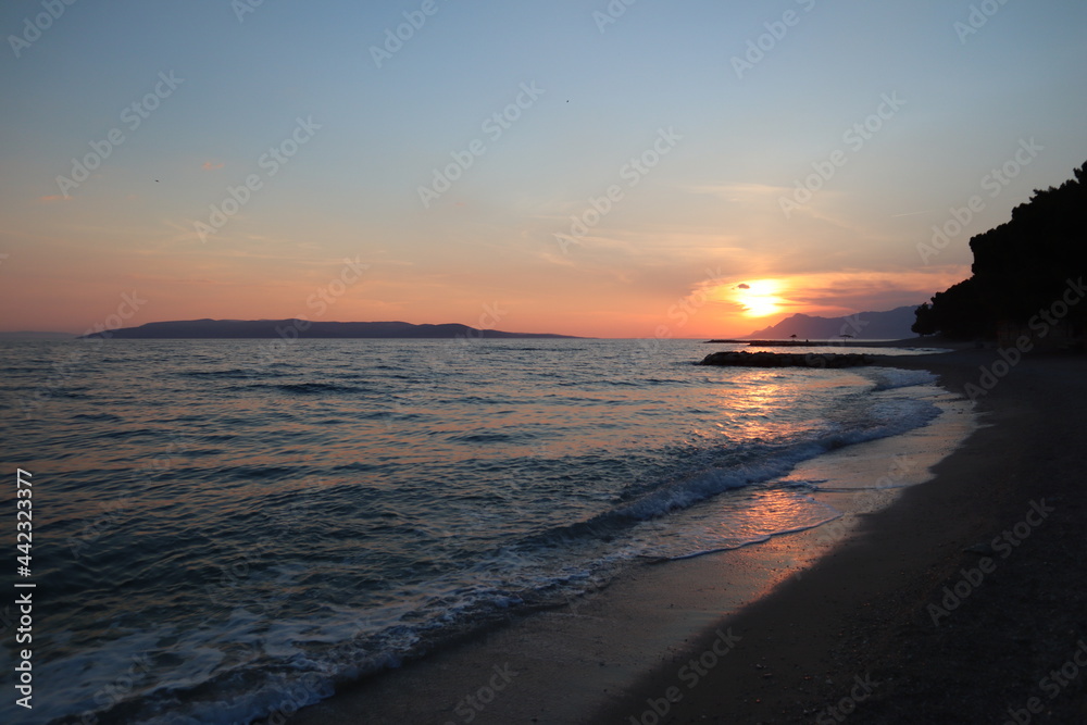 Scenic sunset on the beach on the Croatian seaside.  Seascape evening sea and yellow sun on the horizon, sun glare on the sea surface, rocks and sea foam on a summer evening
