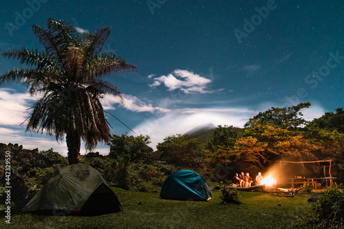 A young group of friends gather around a camp fire at the skirts of a volcano and surrounded by tropical nature.