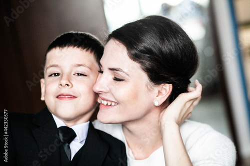 A boy in a school uniform presses his face to his mother and they smile, mother in a white jacket, front view, close-up, daylight