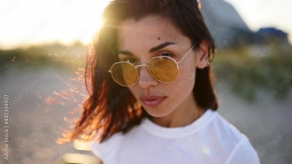 Portrait of young woman looking at camera standing on the beach with a sea on background. Attractive girl in white t-shirt and sunglasses raises eyes. Wind blows airy brown hair shining in sun flares. Portrait of young woman looking at camera standing on the beach with a sea on background. Attractive girl in white t-shirt and sunglasses raises eyes. Wind blows airy brown hair shining in sun flares.