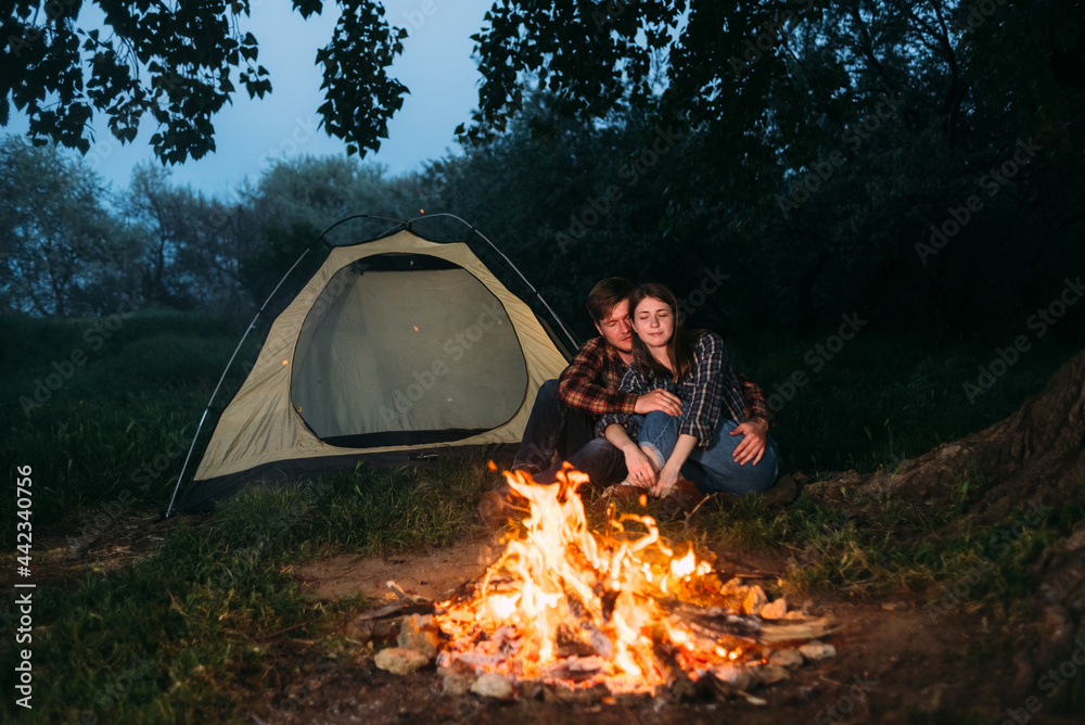 A young couple are sitting near a tent in the forest and making a bonfire in the evening. The branches of the tree are burning. people bask in the hike. Camping guy and girl are resting