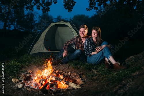 Happy hikers burn a fire in the forest and relax while sitting by the tent in the evening. Young family on vacation. Outdoors. trekking tourism concept.