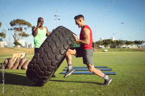 Diverse fit man and trainer exercising outdoors, encouraging and lifting heavy tyre
