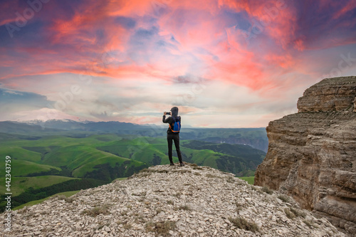 A girl stands on the edge of a cliff and takes pictures of a beautiful view. View from the back. Rest in the mountains. The concept of active recreation. Tourism in the highlands. Rest in solitude.