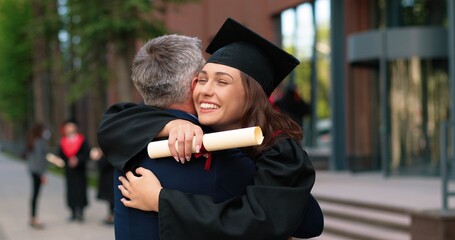 Happy female graduate are embracing with her father with diploma at her hands and rejoicing with each other. Man is hugging his daughter with other students moving and talking in background