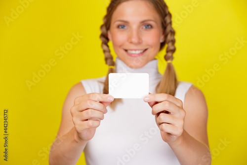 Portrait of pretty and smiling young woman with pigtails holding empty business card isolated on yellow background
