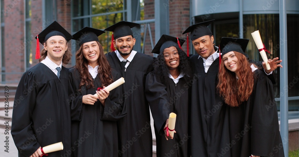 Company of happy multiracial student people are celebrating their graduation while standing near the college and rejoicing with diplomas at the hands. Education concept