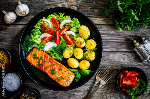 Fried salmon steak with roast potatoes and vegetable salad served on black plate on wooden table
