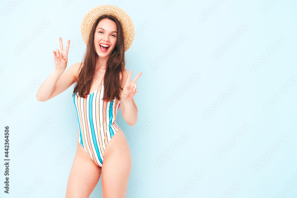 Young beautiful sexy smiling  hipster woman in posing near light blue wall in studio. Trendy model in colorful summer swimwear bathing suit. Positive model shows peace sign.Happy and cheerful in hat