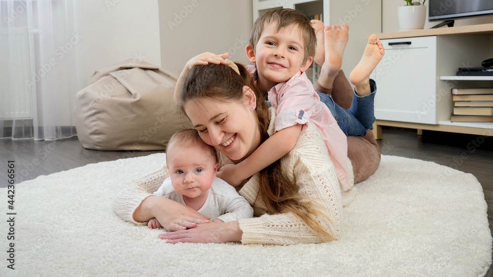 Cute boy hugging his mother and little baby brother lying on carpet in living room. Parenting, children happiness and family relationship