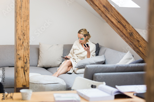 Young female entrepreneur wearing beauty face mask and cosy warm bathrobe in the morning, working remotly from her living room sofa. Remote work from home concept. photo