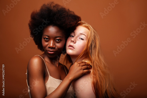 young pretty caucasian, afro woman posing cheerful together on brown background, lifestyle diverse nationality people concept