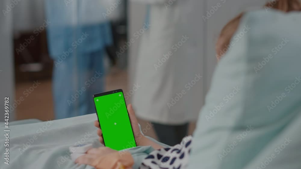 Hospitalized little child resting in bed looking at mock up green screen chroma key smartphone with isolated display. Sick kid suffering medical surgery during recovery consultation in hospital ward Hospitalized little child resting in bed looking at mock up green screen chroma key smartphone with isolated display. Sick kid suffering medical surgery during recovery consultation in hospital ward