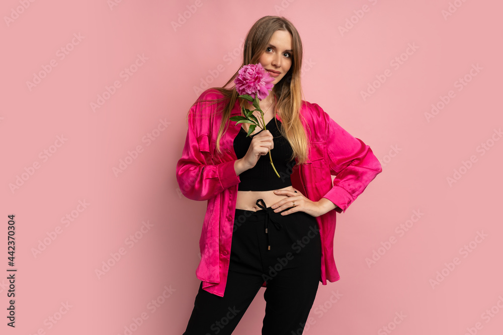 Fashion studio photo of pretty blond woman posing with pion flower in stylish summer outfit over pink background. Fashion studio photo of pretty blond woman posing with pion flower in stylish summer outfit over pink background.