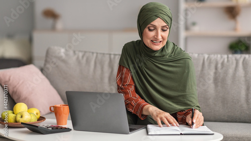 Happy Mature Islamic Lady Using Laptop Taking Notes At Home