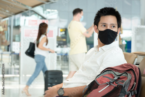 Asian traveler man wearing face mask waiting to board into airplane, standing in departure terminal in airport. Male passenger traveling by plane transportation during covid19 virus pandemic