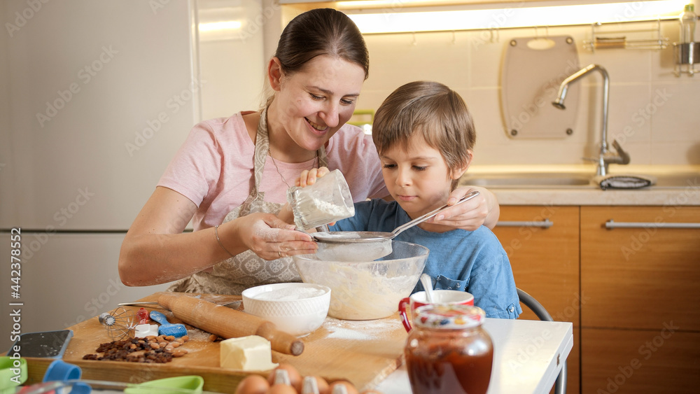 Young smiling mother teaching her son sifting flour with sieve for making biscuit dough. Children cooking with parents, little chef, family having time together, domestic kitchen. Young smiling mother teaching her son sifting flour with sieve for making biscuit dough. Children cooking with parents, little chef, family having time together, domestic kitchen.